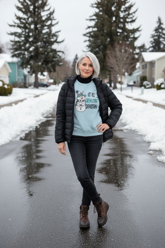 Freezin' Season Sweatshirt, light blue with a snowman and winter lettering, worn under a black jacket.
